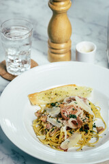 Delicious spaghetti with garnishes on a plate with garlic bread. The dish is presented near a glass of water and a pepper shaker