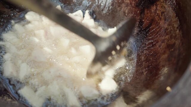 In slow motion, a slotted spoon stirs melting lamb tail fat in a hot cauldron outdoors during the first stage of traditional pilaf preparation, as the fat renders and bubbles in the heat
