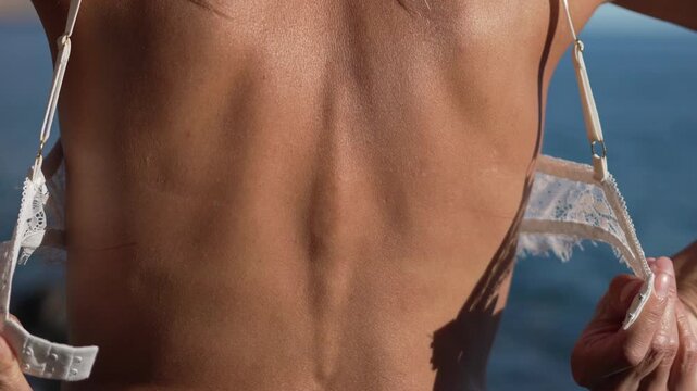 Undressing back bra woman unhooking white lace lingerie outdoors, symbolizing summer freedom and relaxation near the ocean.