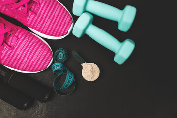 Top view of whey protein powder scoop, teal dumbbells, pink sneakers, jump rope and measuring tape on dark background with copy space, fitness equipment and sports nutrition concept