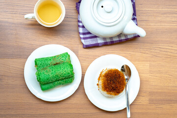 A top-down flat lay of Indonesian snacks, Kue Dadar Gulung and Ketan Serundeng, served with a white teapot and a cup of tea on a wooden table.