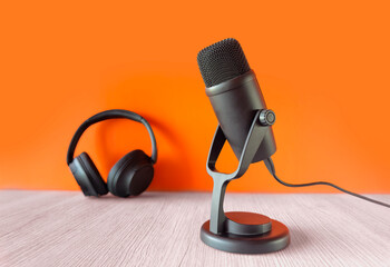 Black microphone and headphones on a light wooden table with an orange background, symbolizing podcasting or recording setup