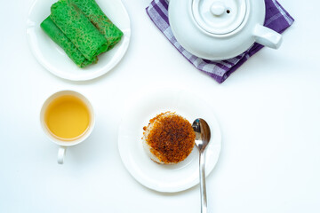 A top-down flat lay of Indonesian snacks, green Kue Dadar Gulung and Ketan Serundeng, served with a cup of tea and a white teapot on a white background.