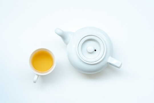 A top-down view of a white teacup filled with tea, placed next to a white teapot on a plain white background.