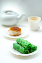 Three green rolled crepes, Kue Dadar Gulung, are in focus on a white plate. In the blurred background are Ketan Serundeng, a cup of tea, and a teapot on a white surface.