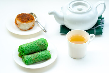 Green rolled crepes, Kue Dadar Gulung, are in focus on a white plate, with a cup of tea, a teapot, and Ketan Serundeng blurred behind them on a white surface.