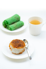 etan Serundeng (sticky rice with toasted coconut) on a white plate, with green Kue Dadar Gulung (rolled crepes) and a cup of tea blurred in the background, all on a white surface.