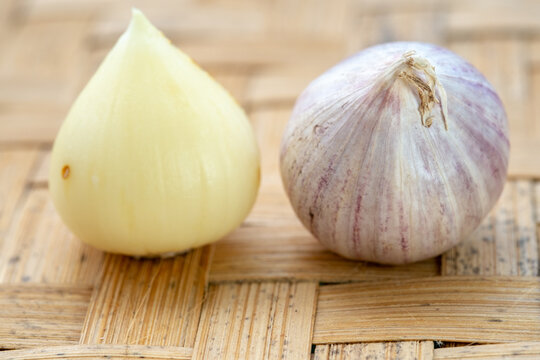 A single peeled solo garlic clove and a whole garlic bulb sitting side-by-side on a traditional woven bamboo tray.