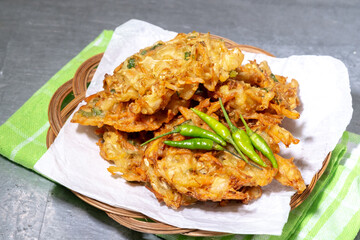 A stack of crispy Indonesian bakwan sayur (vegetable fritters) served on white paper with a group of green chili peppers on a woven bamboo plate.