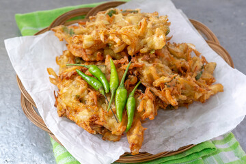 A stack of crispy Indonesian bakwan sayur (vegetable fritters) served on white paper with a group of green chili peppers on a woven bamboo plate.