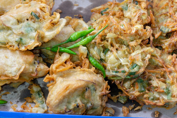 A close-up of Indonesian fried snacks, bakwan (vegetable fritters) and tahu isi (stuffed tofu), piled together with small green chilies on top.