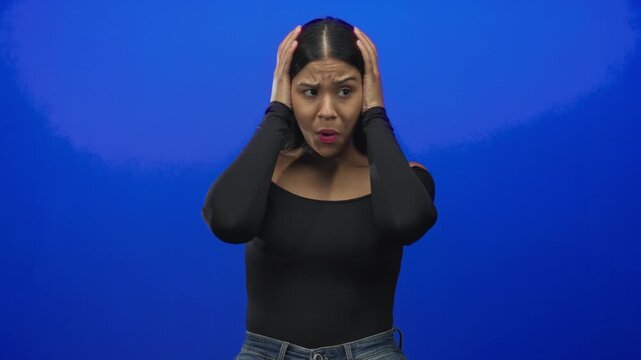 Young latin woman grasping head with both hands in vibrant blue studio wearing black offshoulder top and denim jeans; anxiety.