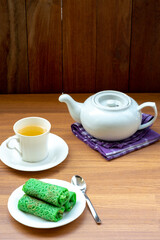 Fototapeta premium A plate of green dadar gulung, Indonesian crepes, served with a cup of tea and a white teapot on a wooden table. A dark wood wall is in the background.