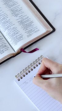 Human hand is writing inspirational biblical quote in a notebook with holy bible on the table. Vertical video. Christian study and education concept.