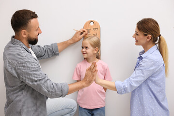 Parents measuring height of their little daughter with scale near white wall indoors