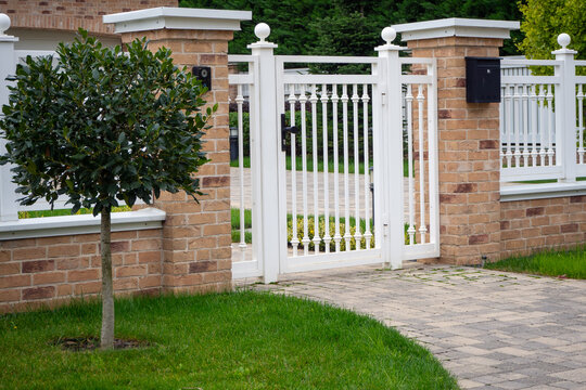 White picket gate between brick pillars at residential entrance. Clean design with green lawn and paved pathway for suburban property. Real photo