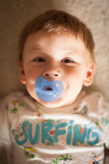 A cheerful toddler lies on a soft surface, wearing a colorful surfing shirt and a blue pacifier. The warm light highlights his joyful expression in a cozy room.