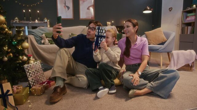 Happy Caucasian family connecting virtually with relatives on Christmas Eve, sitting on cozy rug near decorated tree, smiling and demonstrating present boxes during joyful video chat on smartphone