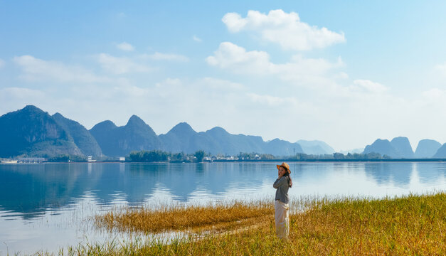 Chinese woman enjoying Quyang Lake (Quyanghu)  karst peaks landscape standing on grassland along lakeside in winter in Jingxi county, Baise city, Guangxi province, China - Powered by Adobe
