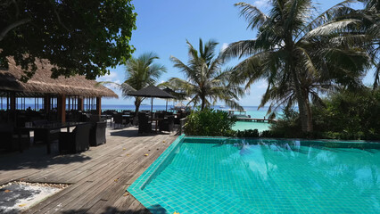 Luxury resort photo of Maldives tropical paradise island in Indian Ocean with hotel territory, palm trees, beach umbrella and chair, swimming pool. Beautiful photography, azure ocean, sky with clouds.
