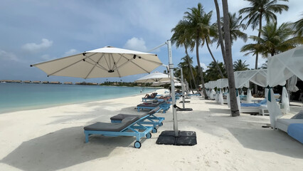 Luxury resort photo of Maldives tropical paradise island in Indian Ocean with hotel territory, palm trees, beach umbrella and chair. Beautiful photography, azure ocean, sky with clouds.
