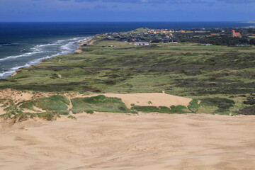 Landscape photo with a view of sand dunes, green hills and the town of Lonstrup in the background near the Rubjerg Knude lighthouse in Denmark