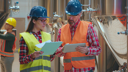 Industrial engineers in hard hats and safety vests are collaborating on a production plan, reviewing data on a digital tablet and clipboard in a modern manufacturing facility