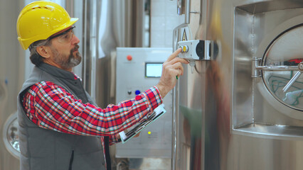Experienced engineer wearing hard hat and safety glasses operating control panel on stainless steel tank, checking production process in modern industrial facility
