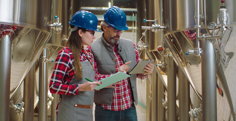 Two brewery workers wearing safety hats and plaid shirts, checking production data on a digital tablet and clipboard next to large stainless steel fermentation tanks