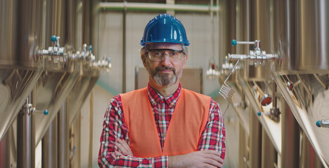 Mature man, wearing a blue hard hat, safety glasses, and an orange vest, standing confidently with arms crossed in a modern brewery production area with stainless steel tanks
