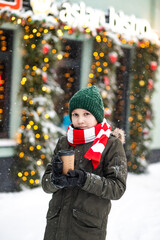 Cute teenager boy kid with a paper cup of hot cocoa or tea in winter on the background of the snow city