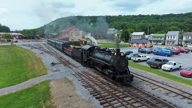 A vintage steam train pulls out of the station, chugging down the tracks in a small town surrounded by green hills. The scene captures the charm of rail travel and the beauty of the area.