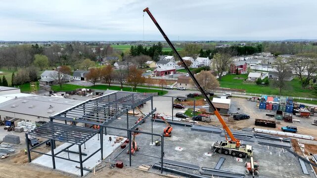 Large construction site shows a crane lifting steel beams into place. Workers coordinate below as structures take shape in the countryside. Green fields surround the area.
