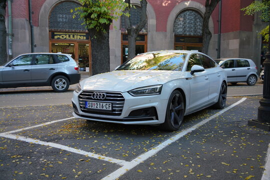 White Audi A5 Sportback (B9 generation) is parked in front of a building with arches. The ground is covered with fallen yellow leaves, the autumn season. Subotica, Serbia, October 22, 2024