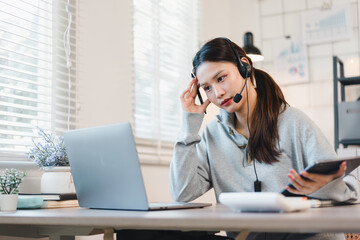 Young woman with headset working at laptop in bright home office, focused and slightly stressed while holding tablet