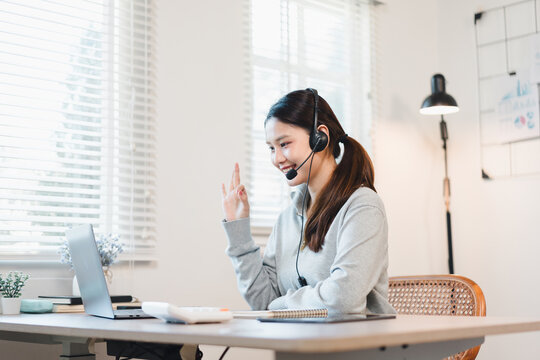 Young woman video call tutoring session at home smiling and waving while wearing headset and sitting at desk with laptop and notebook