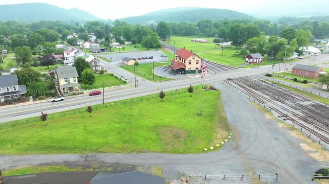 The camera captures a stunning aerial view of a small rail road town featuring green fields and nearby mountains. Roads and buildings are visible, alongside a railway track running through the area.