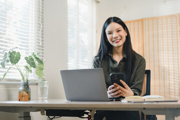 Young woman smiling while working at laptop and holding smartphone in bright home office with plant and notebook