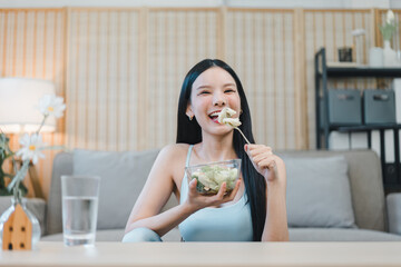Young woman eating salad at home smiling happily with relaxed living room background