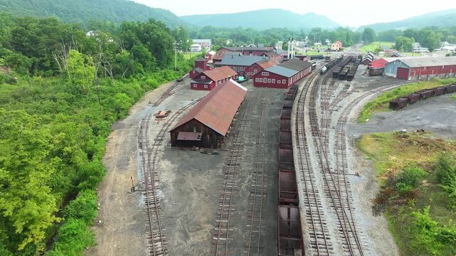 An aerial view showcases an abandoned train yard nestled in a picturesque mountain town. Rusting train cars line the tracks while greenery surrounds the area, adding to its haunting beauty.