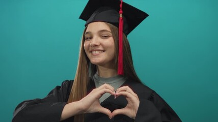Girl wearing graduation cap makes heart gesture in studio with turquoise background; pride success joy.