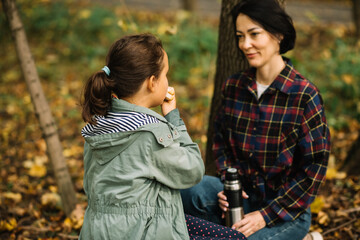 Mother with kid child girl hiking in autumn forest relax sitting on stump, drinks from thermos and eating sandwich in autumn forest.