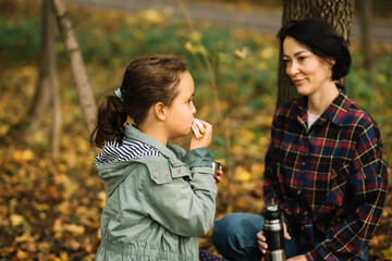 Mother with kid child girl hiking in autumn forest relax sitting on stump, drinks from thermos and eating sandwich in autumn forest.