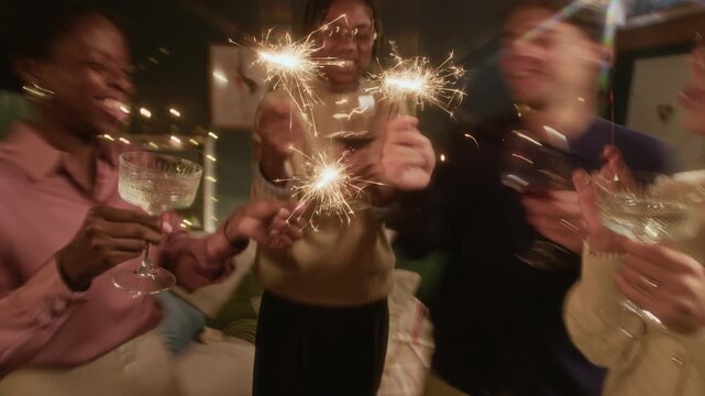 Motion blur slowmo shot of four multiethnic friends with sparklers and glasses in hands partying together during Christmas celebration in cozy home setting with glowing Christmas tree