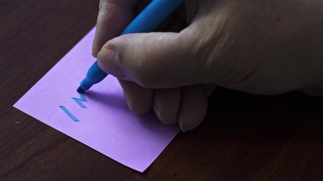 A hand writes text I miss You on a small purple note with a blue marker 