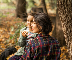Mother with kid child girl hiking in autumn forest relax sitting on stump, drinks from thermos and eating sandwich in autumn forest.