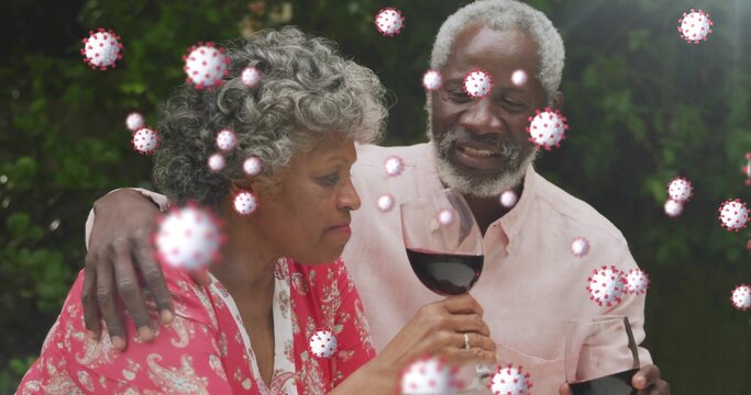 Holding wine glasses senior couple hugging in pink attire at garden table, with virus graphics - Powered by Adobe