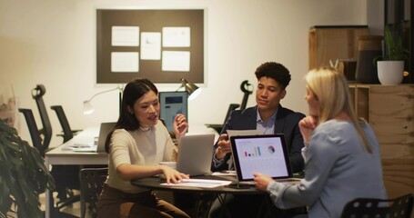 Analyzing three professionals in business attire discussing charts on office table with laptops