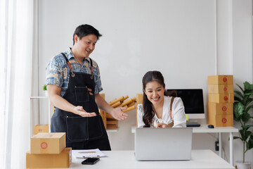 Happy Asian couple entrepreneurs collaborating, checking laptop for e commerce orders, processing packages for delivery in home office