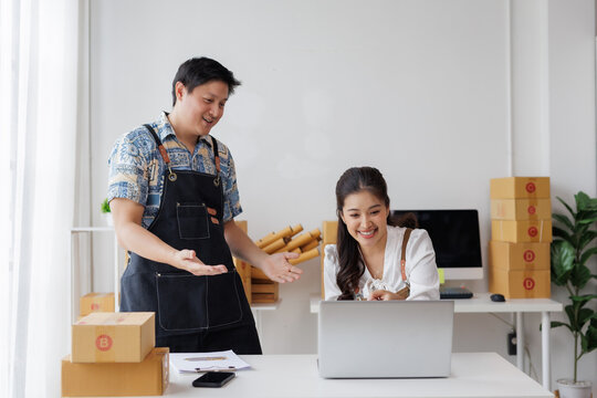 Happy Asian couple entrepreneurs collaborating, checking laptop for e commerce orders, processing packages for delivery in home office